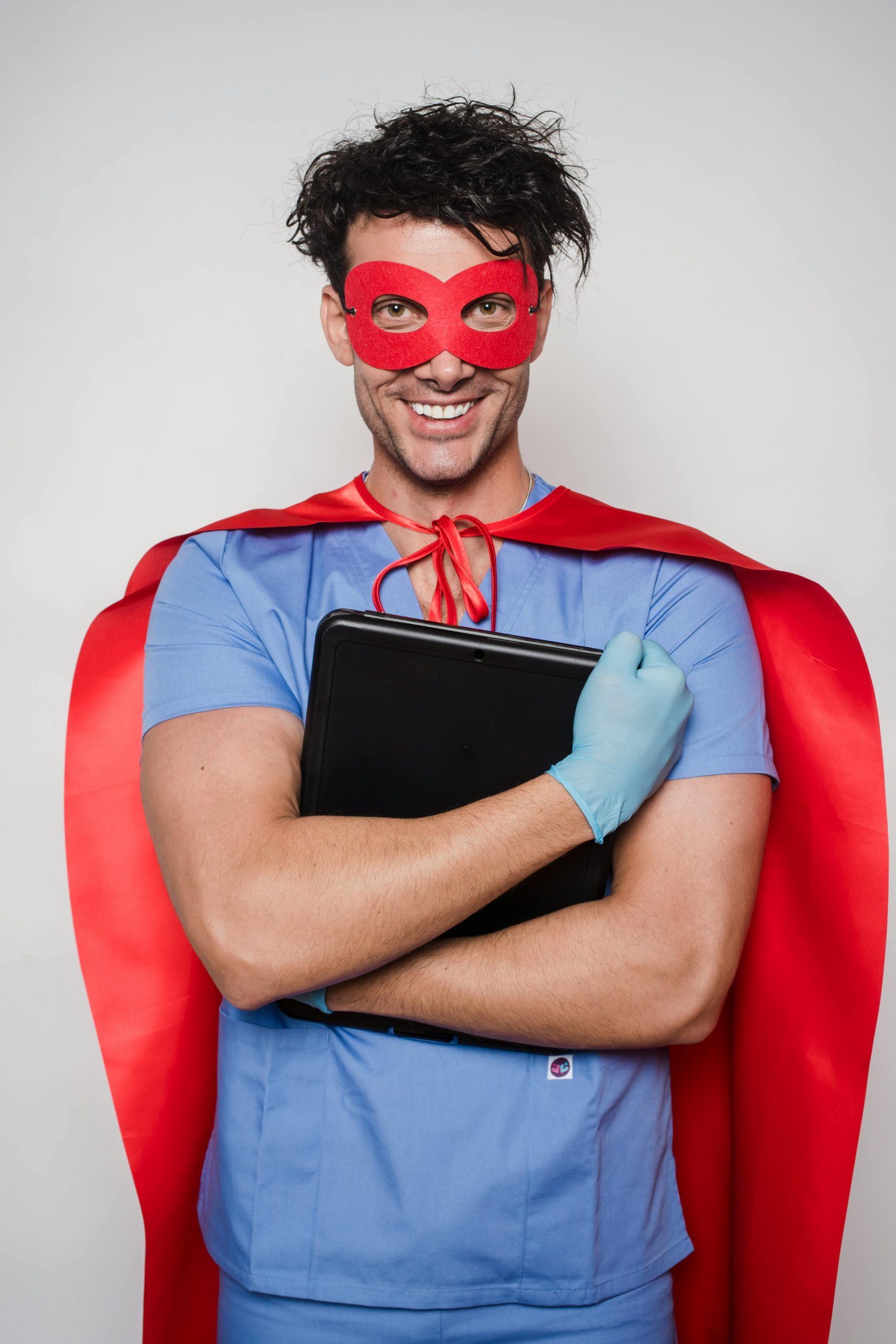Cheerful man doctor in superhero costume with mask and cape standing with folder with documents and looking at camera against light wall